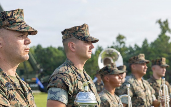 A group of U.S. Marines in digital camouflage (MARPAT) uniforms are standing at attention during a formal military ceremony on a grass field. The foreground Marine wears white ceremonial gloves and holds a mace or ceremonial staff, while behind him several Marines hold brass instruments including trumpets and a saxophone. An American flag is partially visible in the background along with trees and military equipment.