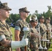 A group of U.S. Marines in digital camouflage (MARPAT) uniforms are standing at attention during a formal military ceremony on a grass field. The foreground Marine wears white ceremonial gloves and holds a mace or ceremonial staff, while behind him several Marines hold brass instruments including trumpets and a saxophone. An American flag is partially visible in the background along with trees and military equipment.