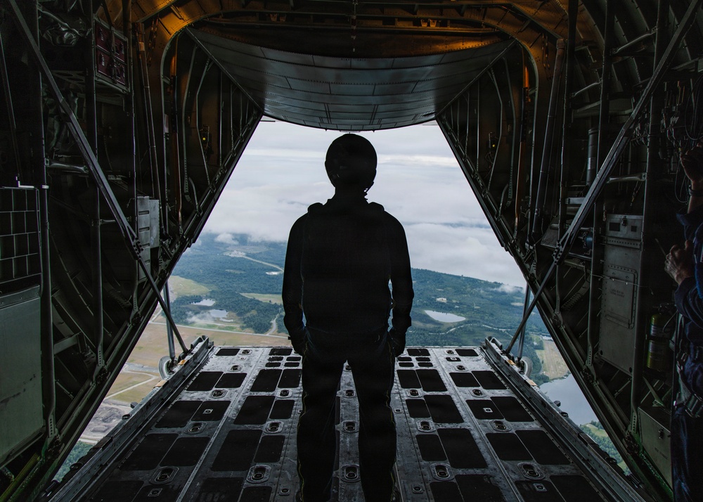 A silhouetted figure stands at the open rear cargo ramp of a military transport aircraft in flight, looking out over a forested landscape with scattered clouds and bodies of water below. The interior of the aircraft shows typical military cargo hold features including structural framing, tie-down rings, and equipment panels. The dramatic backlighting creates a strong silhouette effect against the aerial landscape.