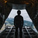 A silhouetted figure stands at the open rear cargo ramp of a military transport aircraft in flight, looking out over a forested landscape with scattered clouds and bodies of water below. The interior of the aircraft shows typical military cargo hold features including structural framing, tie-down rings, and equipment panels. The dramatic backlighting creates a strong silhouette effect against the aerial landscape.