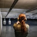 A person with red hair in a ponytail is standing at an indoor shooting range, aiming a handgun downrange at silhouette targets. They are wearing full multicam camouflage uniform, ear protection (earmuffs), and safety glasses. Spent brass casings are visible on the floor near a equipment tray to the left.
