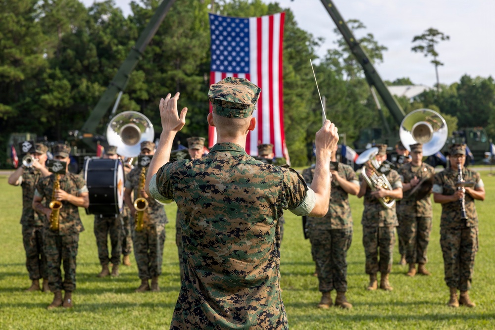 A U.S. Marine Corps band performs outdoors on a grass field, led by a conductor in digital camouflage uniform holding a baton. A large American flag is prominently displayed in the background, suspended between two military cranes or booms. The setting appears to be a formal military ceremony or parade.
