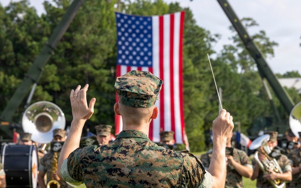 A U.S. Marine Corps band performs outdoors on a grass field, led by a conductor in digital camouflage uniform holding a baton. A large American flag is prominently displayed in the background, suspended between two military cranes or booms. The setting appears to be a formal military ceremony or parade.