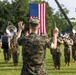 A U.S. Marine Corps band performs outdoors on a grass field, led by a conductor in digital camouflage uniform holding a baton. A large American flag is prominently displayed in the background, suspended between two military cranes or booms. The setting appears to be a formal military ceremony or parade.
