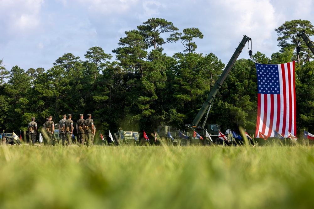 A military ceremony or event is taking place on an open grassy field, featuring uniformed personnel standing in formation alongside military vehicles. A large American flag is prominently displayed, suspended from what appears to be a military crane or boom arm. Multiple state flags are also visible in a row, suggesting a formal patriotic or commemorative event.