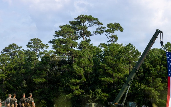A military ceremony or event is taking place on an open grassy field, featuring uniformed personnel standing in formation alongside military vehicles. A large American flag is prominently displayed, suspended from what appears to be a military crane or boom arm. Multiple state flags are also visible in a row, suggesting a formal patriotic or commemorative event.