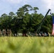 A military ceremony or event is taking place on an open grassy field, featuring uniformed personnel standing in formation alongside military vehicles. A large American flag is prominently displayed, suspended from what appears to be a military crane or boom arm. Multiple state flags are also visible in a row, suggesting a formal patriotic or commemorative event.