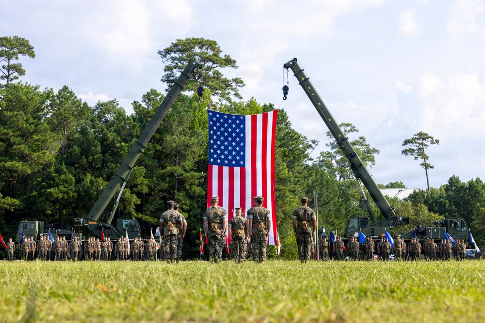 A formal military ceremony is taking place on an open grass field, featuring a large American flag suspended between two military cranes. Personnel in camouflage uniforms are assembled in formation on both sides, with a small group of individuals standing at attention directly in front of the flag. The setting appears to be a military installation surrounded by trees.