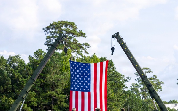 A formal military ceremony is taking place on an open grass field, featuring a large American flag suspended between two military cranes. Personnel in camouflage uniforms are assembled in formation on both sides, with a small group of individuals standing at attention directly in front of the flag. The setting appears to be a military installation surrounded by trees.