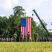 A formal military ceremony is taking place on an open grass field, featuring a large American flag suspended between two military cranes. Personnel in camouflage uniforms are assembled in formation on both sides, with a small group of individuals standing at attention directly in front of the flag. The setting appears to be a military installation surrounded by trees.
