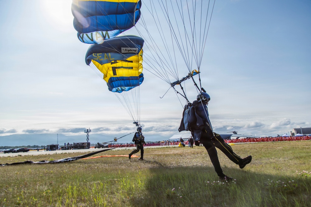 Two skydivers are landing at what appears to be a military airshow, with blue and yellow parachutes prominently displaying the word 'SEAL' and 'NAVY', indicating this is a U.S. Navy SEAL parachute demonstration team. The jumpers are touching down on a grassy airfield with a large crowd of spectators visible in the background. Aircraft hangars and ground support equipment are visible on the airfield.