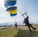 Two skydivers are landing at what appears to be a military airshow, with blue and yellow parachutes prominently displaying the word 'SEAL' and 'NAVY', indicating this is a U.S. Navy SEAL parachute demonstration team. The jumpers are touching down on a grassy airfield with a large crowd of spectators visible in the background. Aircraft hangars and ground support equipment are visible on the airfield.