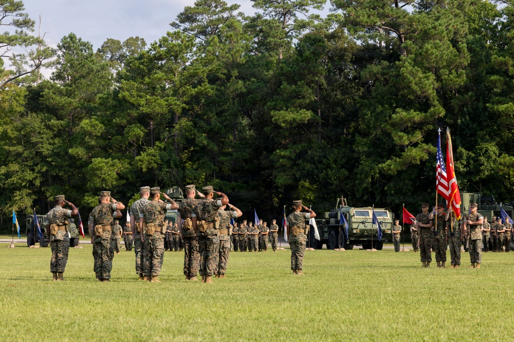 A formal U.S. Marine Corps military ceremony is taking place on an outdoor parade field surrounded by trees. Multiple Marines in woodland MARPAT camouflage uniforms are rendering salutes while others hold the U.S. flag and Marine Corps colors. Military vehicles, likely MRAPs or similar tactical vehicles, are positioned in the background as part of the formation.