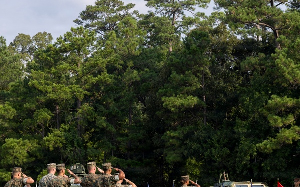 A formal U.S. Marine Corps military ceremony is taking place on an outdoor parade field surrounded by trees. Multiple Marines in woodland MARPAT camouflage uniforms are rendering salutes while others hold the U.S. flag and Marine Corps colors. Military vehicles, likely MRAPs or similar tactical vehicles, are positioned in the background as part of the formation.