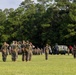 A formal U.S. Marine Corps military ceremony is taking place on an outdoor parade field surrounded by trees. Multiple Marines in woodland MARPAT camouflage uniforms are rendering salutes while others hold the U.S. flag and Marine Corps colors. Military vehicles, likely MRAPs or similar tactical vehicles, are positioned in the background as part of the formation.