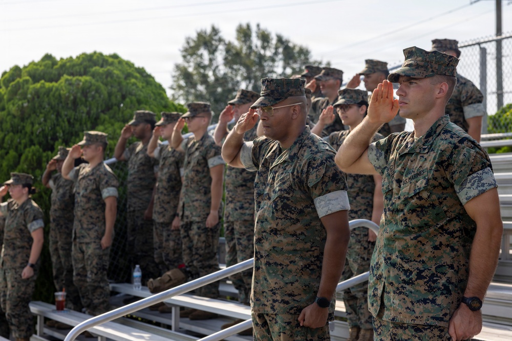 A group of U.S. Marines in MARPAT (Marine Pattern) camouflage uniforms are standing on bleachers rendering a salute during what appears to be a formal military ceremony or event. The personnel are arranged in rows on stadium-style bleachers, all facing the same direction with right hands raised in salute. The setting appears to be an outdoor athletic or parade field during daylight hours.