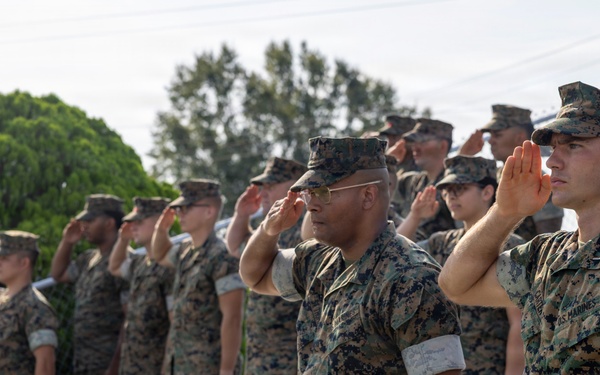 A group of U.S. Marines in MARPAT (Marine Pattern) camouflage uniforms are standing on bleachers rendering a salute during what appears to be a formal military ceremony or event. The personnel are arranged in rows on stadium-style bleachers, all facing the same direction with right hands raised in salute. The setting appears to be an outdoor athletic or parade field during daylight hours.