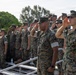 A group of U.S. Marines in MARPAT (Marine Pattern) camouflage uniforms are standing on bleachers rendering a salute during what appears to be a formal military ceremony or event. The personnel are arranged in rows on stadium-style bleachers, all facing the same direction with right hands raised in salute. The setting appears to be an outdoor athletic or parade field during daylight hours.