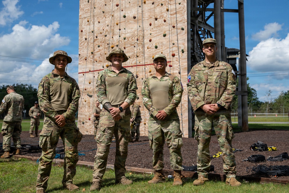 Four military personnel in OCP (Operational Camouflage Pattern) uniforms pose together in front of a large wooden climbing wall at what appears to be a military training facility. The rightmost soldier displays a patch consistent with the 82nd Airborne Division on their left shoulder. The setting suggests an outdoor obstacle course or physical training area.