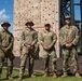 Four military personnel in OCP (Operational Camouflage Pattern) uniforms pose together in front of a large wooden climbing wall at what appears to be a military training facility. The rightmost soldier displays a patch consistent with the 82nd Airborne Division on their left shoulder. The setting suggests an outdoor obstacle course or physical training area.