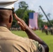 A U.S. Marine in dress uniform is photographed from behind rendering a salute toward a large American flag displayed at an outdoor military ceremony. Rows of uniformed personnel are visible in formation on a green field in the background. The image captures a formal military parade or ceremony setting.