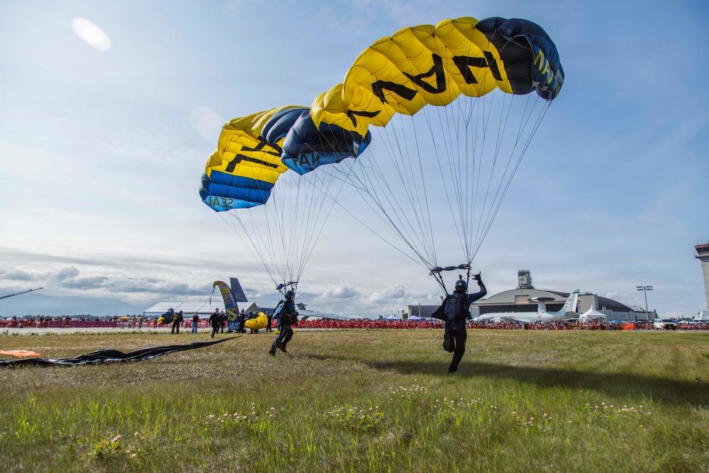 Two military skydivers from the U.S. Navy SEAL parachute demonstration team are landing on a grassy airfield during what appears to be a public air show. The parachutes are distinctively colored in yellow and black with 'NAVY' and 'SEAL' text visible. A large crowd of spectators lines the airfield in the background near military hangars and aircraft.