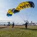 Two military skydivers from the U.S. Navy SEAL parachute demonstration team are landing on a grassy airfield during what appears to be a public air show. The parachutes are distinctively colored in yellow and black with 'NAVY' and 'SEAL' text visible. A large crowd of spectators lines the airfield in the background near military hangars and aircraft.