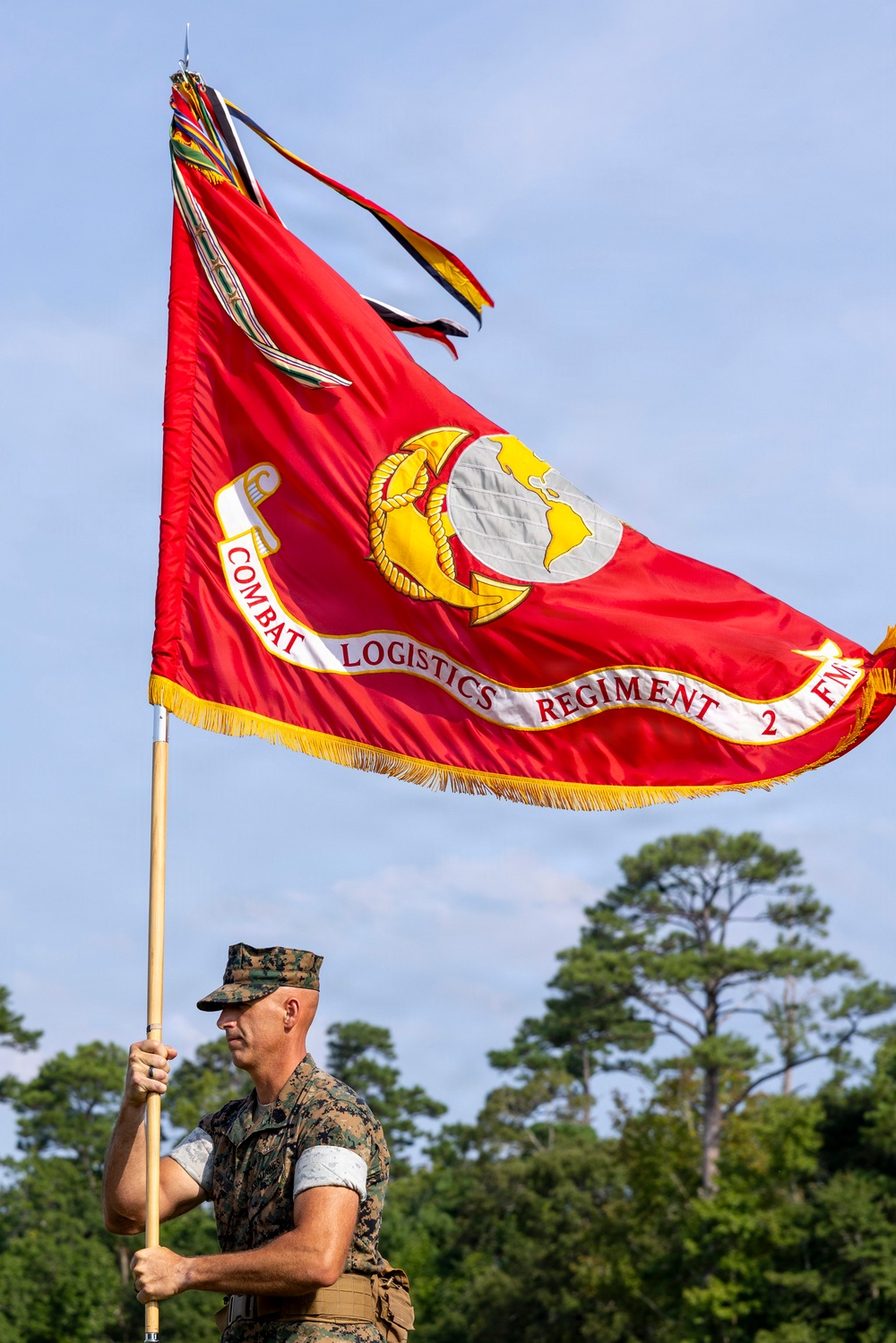 A U.S. Marine in combat utility uniform (MARPAT camouflage) carries the guidon of Combat Logistics Regiment 2, Fleet Marine Corps (FMC), during what appears to be a formal military ceremony. The large red flag features the Marine Corps Eagle, Globe, and Anchor emblem in gold and silver, bordered with gold fringe. The setting appears to be an outdoor parade field with trees visible in the background.