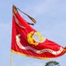 A U.S. Marine in combat utility uniform (MARPAT camouflage) carries the guidon of Combat Logistics Regiment 2, Fleet Marine Corps (FMC), during what appears to be a formal military ceremony. The large red flag features the Marine Corps Eagle, Globe, and Anchor emblem in gold and silver, bordered with gold fringe. The setting appears to be an outdoor parade field with trees visible in the background.