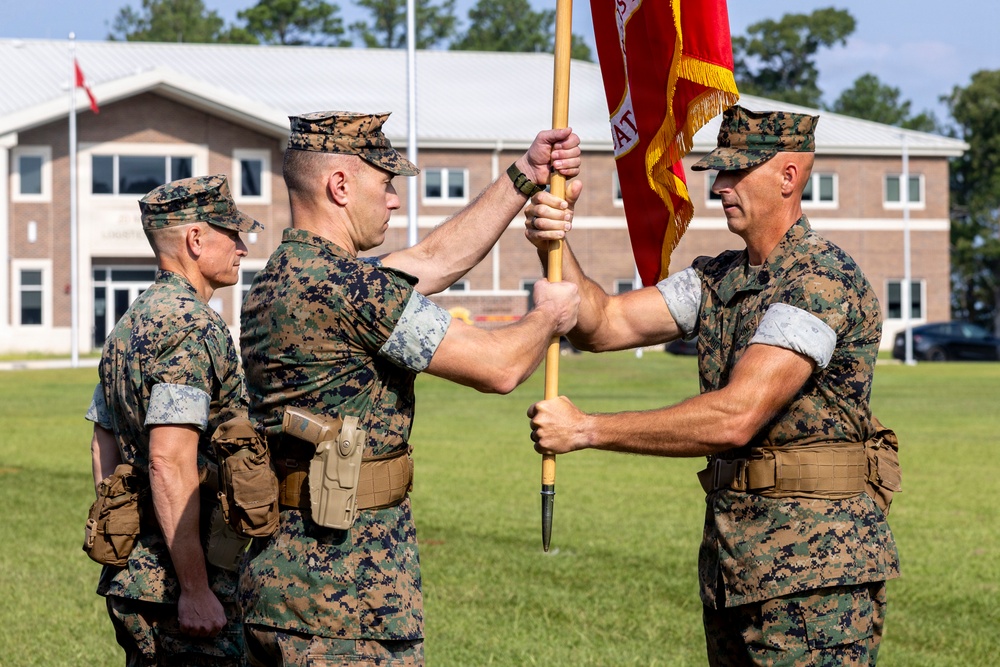 Three U.S. Marine Corps personnel in MARPAT woodland camouflage uniforms are conducting a formal change of command ceremony on a parade field. Two central figures are transferring a red unit guidon with gold fringe, symbolizing the passing of command authority. The ceremony takes place in front of a brick military building on what appears to be a Marine Corps installation.
