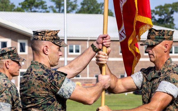 Three U.S. Marine Corps personnel in MARPAT woodland camouflage uniforms are conducting a formal change of command ceremony on a parade field. Two central figures are transferring a red unit guidon with gold fringe, symbolizing the passing of command authority. The ceremony takes place in front of a brick military building on what appears to be a Marine Corps installation.