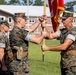 Three U.S. Marine Corps personnel in MARPAT woodland camouflage uniforms are conducting a formal change of command ceremony on a parade field. Two central figures are transferring a red unit guidon with gold fringe, symbolizing the passing of command authority. The ceremony takes place in front of a brick military building on what appears to be a Marine Corps installation.