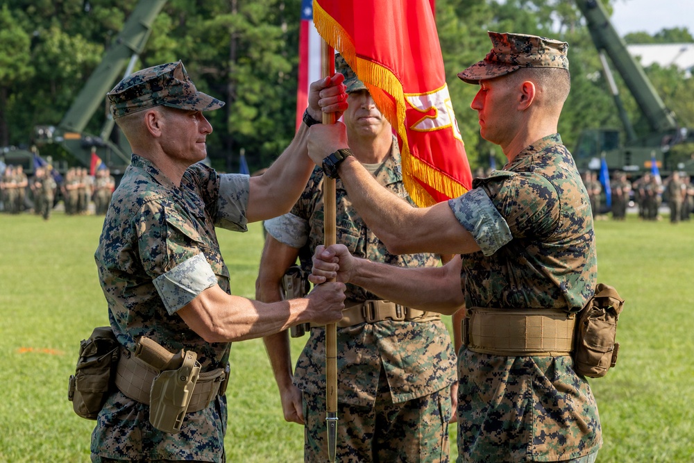 A formal U.S. Marine Corps change of command ceremony is taking place on a parade field. Two Marines in the foreground are exchanging a red unit guidon/colors with gold fringe, while a third Marine holds the staff in the background. Formations of Marines and military equipment are visible in the background.