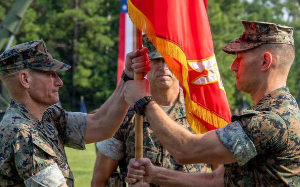 A formal U.S. Marine Corps change of command ceremony is taking place on a parade field. Two Marines in the foreground are exchanging a red unit guidon/colors with gold fringe, while a third Marine holds the staff in the background. Formations of Marines and military equipment are visible in the background.