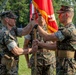 A formal U.S. Marine Corps change of command ceremony is taking place on a parade field. Two Marines in the foreground are exchanging a red unit guidon/colors with gold fringe, while a third Marine holds the staff in the background. Formations of Marines and military equipment are visible in the background.