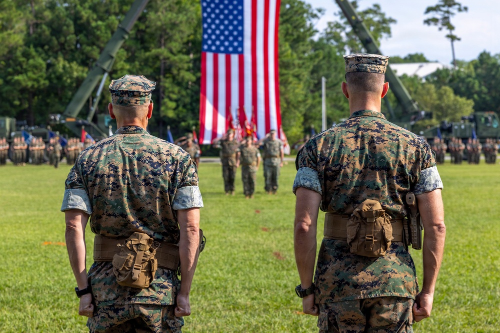 Two U.S. Marines in digital camouflage (MARPAT) uniforms stand at attention on a parade field facing a large American flag during what appears to be a formal military ceremony. A formation of troops and color guard are visible in the background, along with military vehicles and equipment. The setting suggests a change of command, retirement, or unit activation ceremony.
