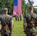 Two U.S. Marines in digital camouflage (MARPAT) uniforms stand at attention on a parade field facing a large American flag during what appears to be a formal military ceremony. A formation of troops and color guard are visible in the background, along with military vehicles and equipment. The setting suggests a change of command, retirement, or unit activation ceremony.