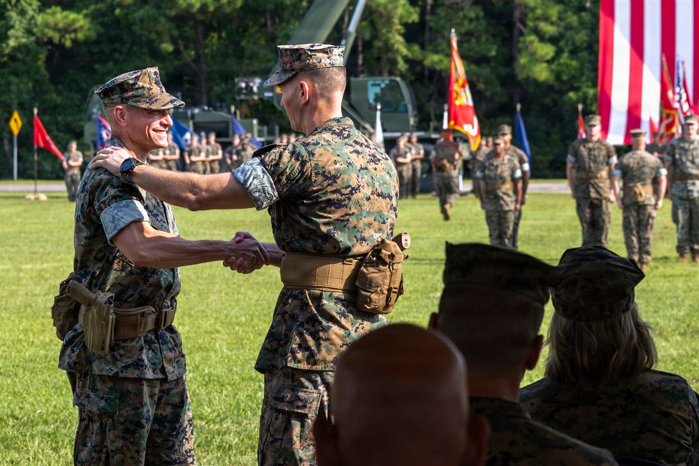 Two U.S. Marine Corps officers are shaking hands during what appears to be a formal military ceremony, likely a change of command or unit activation event. The ceremony is taking place on a grass field with formation troops, color guard, military vehicles, and flags visible in the background. The smiling officer on the left suggests a celebratory or congratulatory moment.