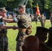 Two U.S. Marine Corps officers are shaking hands during what appears to be a formal military ceremony, likely a change of command or unit activation event. The ceremony is taking place on a grass field with formation troops, color guard, military vehicles, and flags visible in the background. The smiling officer on the left suggests a celebratory or congratulatory moment.