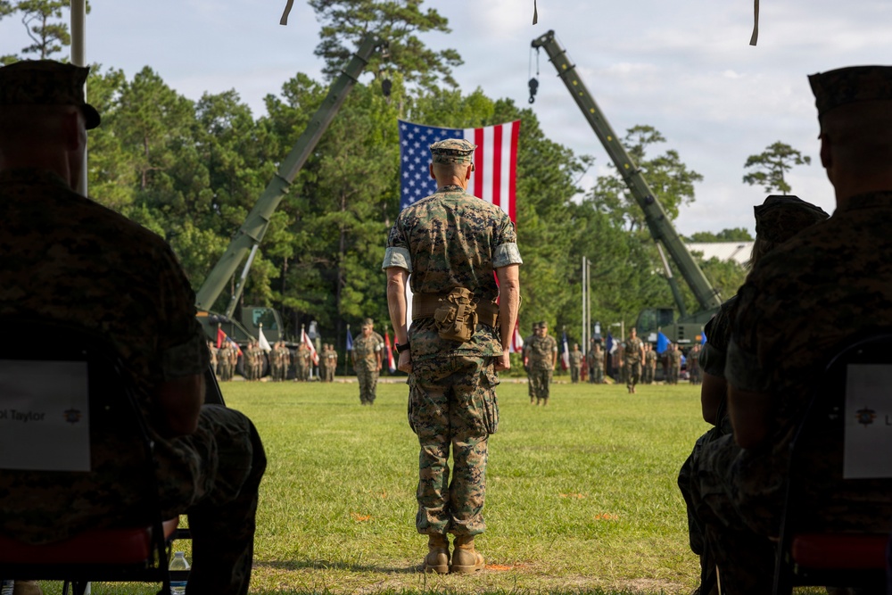 A U.S. Marine Corps military ceremony is taking place on a grass field, with a central figure in MARPAT camouflage uniform standing at attention facing a large American flag suspended between two military cranes. Rows of uniformed Marines are assembled in formation in the background, with additional personnel seated in the foreground. The setting and arrangement suggest a formal change of command or unit ceremony.