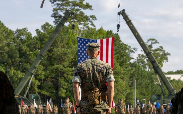A U.S. Marine Corps military ceremony is taking place on a grass field, with a central figure in MARPAT camouflage uniform standing at attention facing a large American flag suspended between two military cranes. Rows of uniformed Marines are assembled in formation in the background, with additional personnel seated in the foreground. The setting and arrangement suggest a formal change of command or unit ceremony.