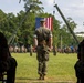 A U.S. Marine Corps military ceremony is taking place on a grass field, with a central figure in MARPAT camouflage uniform standing at attention facing a large American flag suspended between two military cranes. Rows of uniformed Marines are assembled in formation in the background, with additional personnel seated in the foreground. The setting and arrangement suggest a formal change of command or unit ceremony.
