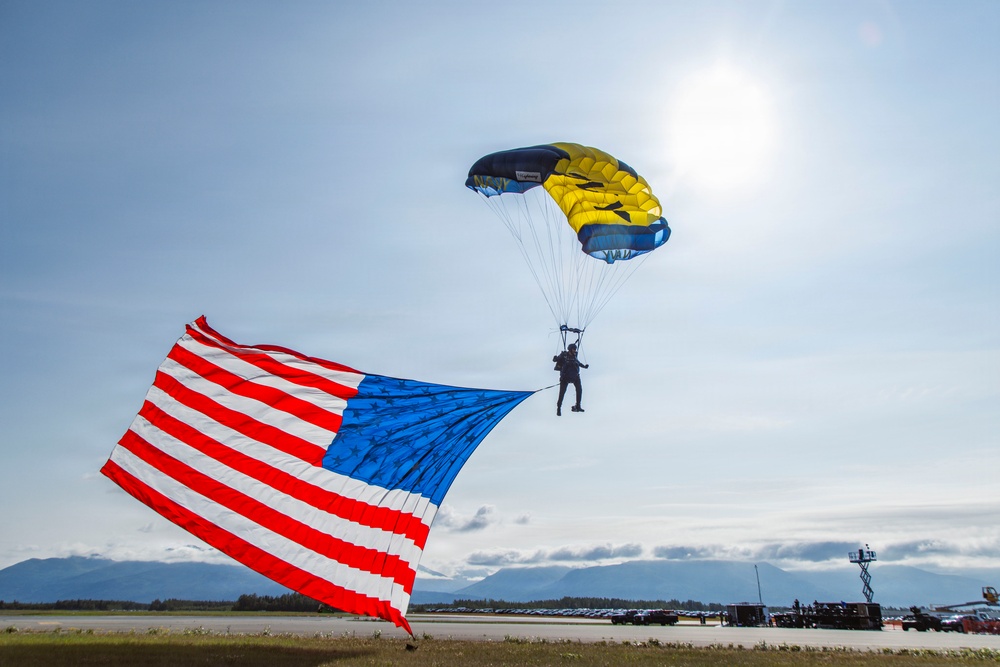 A parachutist is descending toward an airfield runway while carrying a large American flag, which billows dramatically in the wind. The parachute canopy is yellow and dark blue/black in color, and mountains are visible in the background. This appears to be a ceremonial skydiving demonstration, likely at an air show or military event.