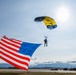 A parachutist is descending toward an airfield runway while carrying a large American flag, which billows dramatically in the wind. The parachute canopy is yellow and dark blue/black in color, and mountains are visible in the background. This appears to be a ceremonial skydiving demonstration, likely at an air show or military event.