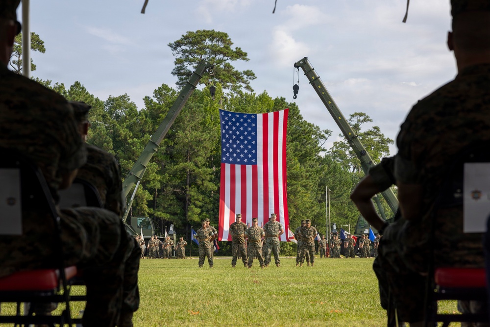 A formal military ceremony is taking place on an open grass field, with a large American flag suspended between two crane arms as a backdrop. Personnel in camouflage uniforms are assembled in formation, with a small group of officers standing at center field facing the seated audience in the foreground. The setting appears to be a change of command or similar official military ceremony at what is likely a U.S. Marine Corps installation.