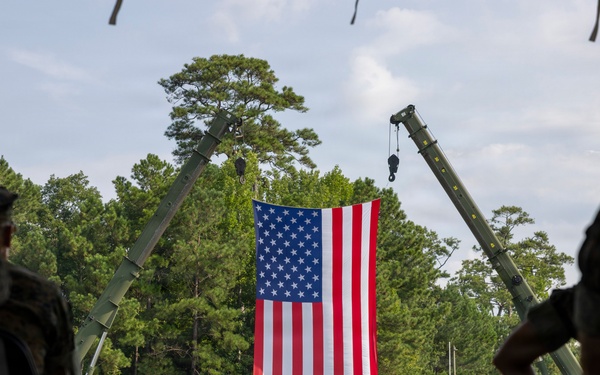A formal military ceremony is taking place on an open grass field, with a large American flag suspended between two crane arms as a backdrop. Personnel in camouflage uniforms are assembled in formation, with a small group of officers standing at center field facing the seated audience in the foreground. The setting appears to be a change of command or similar official military ceremony at what is likely a U.S. Marine Corps installation.