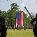 A formal military ceremony is taking place on an open grass field, with a large American flag suspended between two crane arms as a backdrop. Personnel in camouflage uniforms are assembled in formation, with a small group of officers standing at center field facing the seated audience in the foreground. The setting appears to be a change of command or similar official military ceremony at what is likely a U.S. Marine Corps installation.
