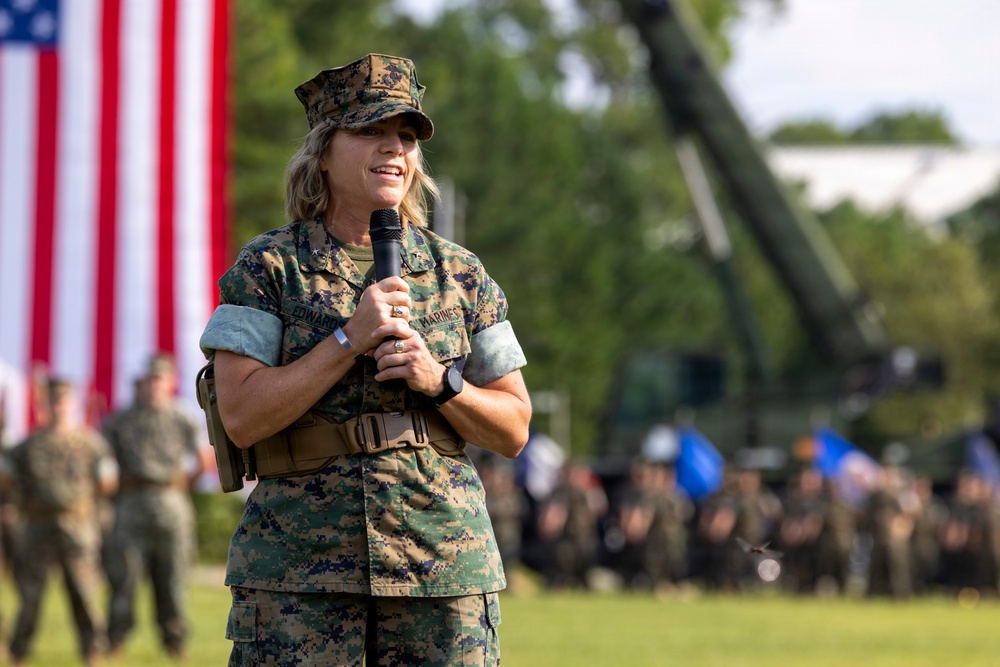 A female U.S. Marine in digital camouflage (MARPAT) uniform is speaking into a microphone at an outdoor military ceremony or formation. A large American flag is prominently displayed in the background, with formations of Marines and military equipment visible behind her. The setting appears to be a formal military event on a parade field.