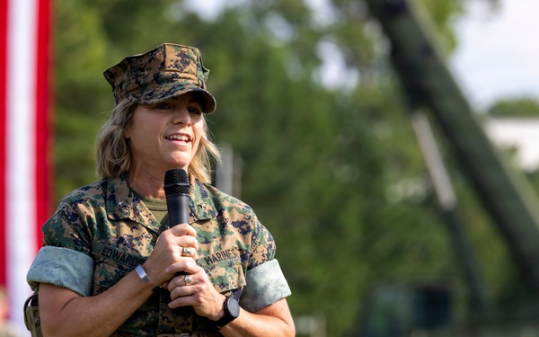 A female U.S. Marine in digital camouflage (MARPAT) uniform is speaking into a microphone at an outdoor military ceremony or formation. A large American flag is prominently displayed in the background, with formations of Marines and military equipment visible behind her. The setting appears to be a formal military event on a parade field.