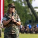A female U.S. Marine in digital camouflage (MARPAT) uniform is speaking into a microphone at an outdoor military ceremony or formation. A large American flag is prominently displayed in the background, with formations of Marines and military equipment visible behind her. The setting appears to be a formal military event on a parade field.